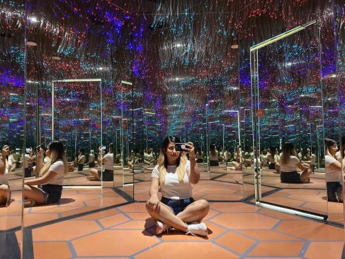 A person sits cross-legged in a mirrored room with colorful lights and reflective surfaces all around.