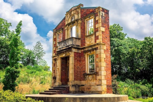 A partially ruined brick building with ornate details, surrounded by greenery and under a cloudy sky.