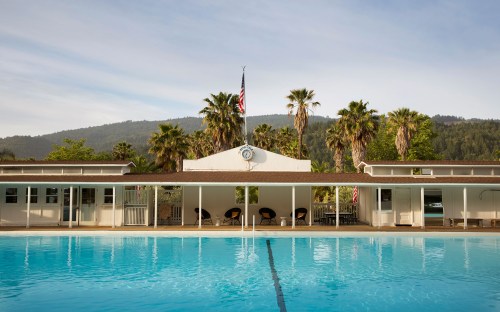 A serene pool scene with palm trees, a building, and a flag against a backdrop of mountains and clear skies.