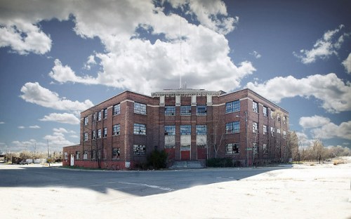 Abandoned brick building with large windows, surrounded by a cloudy sky and empty lot.