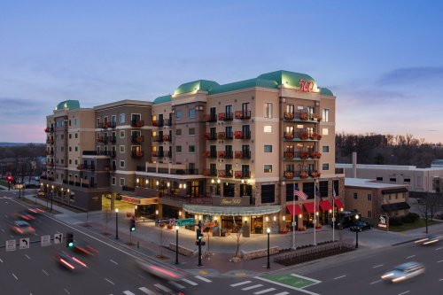 A modern hotel with balconies, illuminated at dusk, located at a busy intersection with flags in front.