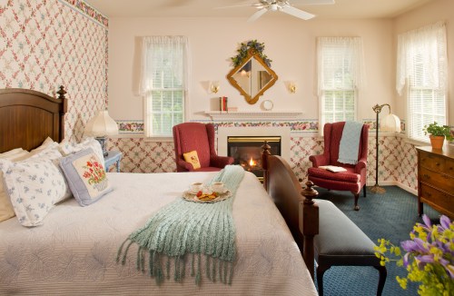 Cozy bedroom with a floral wallpaper, a bed with a tray of snacks, a fireplace, and two armchairs.