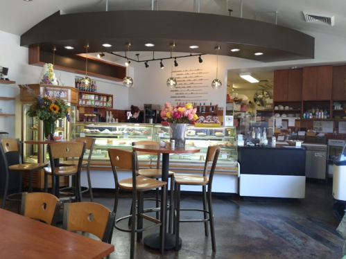 Cozy café interior with a display case of pastries, wooden tables, and colorful flower arrangements.