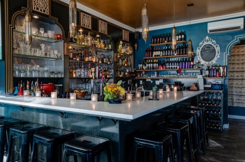 A stylish bar with a sleek white countertop, shelves of colorful bottles, and warm candlelight creating a cozy atmosphere.