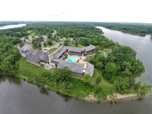 Aerial view of a resort by a river, featuring buildings, a pool, and lush greenery surrounding the area.