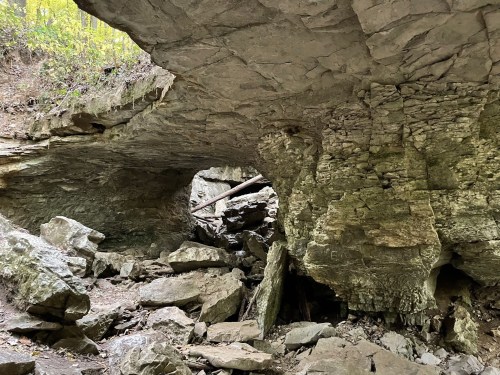 A rocky cave with a large overhanging ledge and scattered boulders, surrounded by greenery.