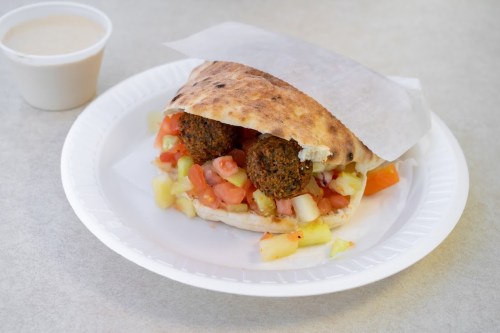 A pita sandwich filled with falafel, diced tomatoes, and cucumbers, served with a small cup of sauce on the side.