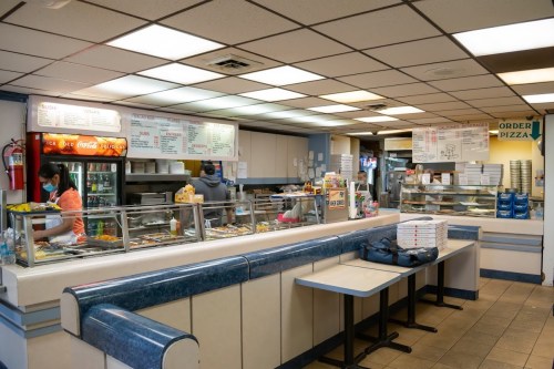 A casual pizza restaurant interior with a counter, menu boards, and staff preparing food. Boxes of pizza are visible.