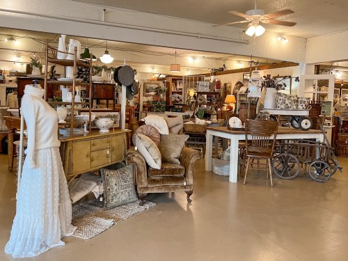 A cozy vintage shop filled with furniture, decor, and a mannequin dressed in a white outfit. Warm, inviting atmosphere.