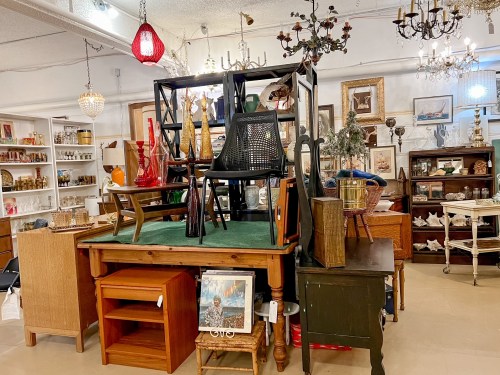 A vintage shop display featuring stacked furniture, including chairs and tables, surrounded by decorative items and artwork.