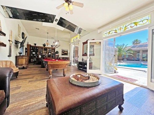 Bright living space with a pool table, chess set, and large windows overlooking a pool and palm trees outside.