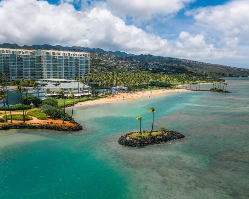 Aerial view of a tropical beach with palm trees, a hotel, and clear turquoise waters under a partly cloudy sky.