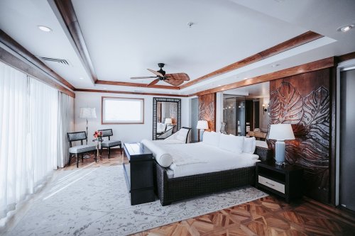 A spacious hotel room featuring a large bed, wooden accents, a seating area, and natural light from large windows.