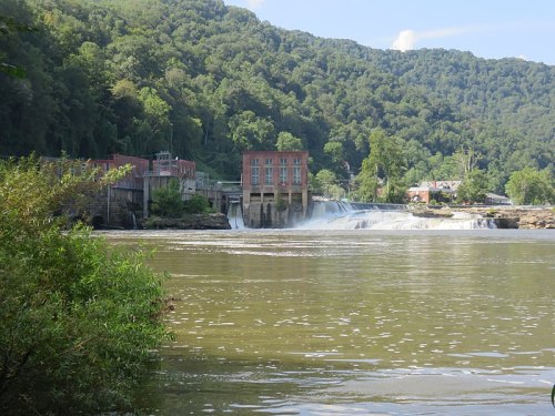 A scenic view of a dam with a waterfall, surrounded by lush green hills and a clear blue sky.