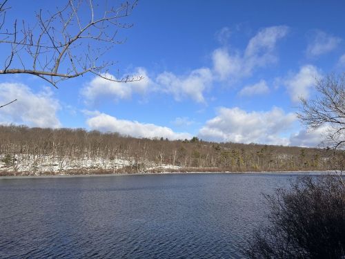 A serene winter landscape featuring a calm lake, snow-dusted trees, and a blue sky with fluffy clouds.