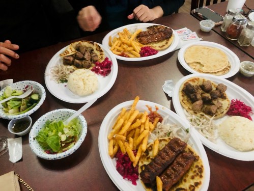 A table with plates of grilled meat, rice, fries, and salad, surrounded by hands reaching for the food.
