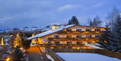 A cozy mountain lodge decorated with lights, surrounded by snow and trees, against a backdrop of mountains at dusk.