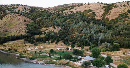 Aerial view of a serene lakeside landscape with hills, trees, and a small campsite featuring tents and cabins.