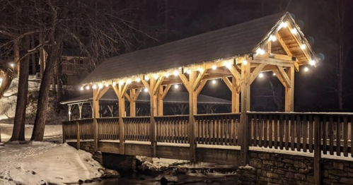 A snow-covered wooden bridge adorned with warm lights, surrounded by trees at night.