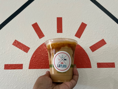 A hand holding a cup of iced coffee with a logo in front of a sunburst mural on a wall.