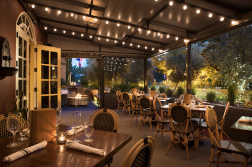 Cozy restaurant patio with string lights, wooden tables, and elegant chairs, surrounded by greenery at dusk.