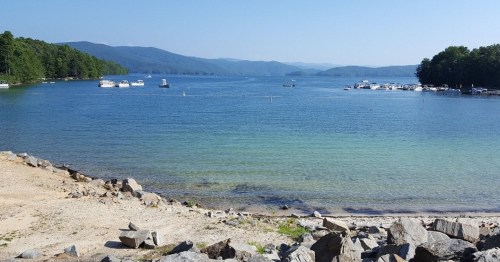 A serene lake scene with boats, rocky shore, and mountains in the background under a clear blue sky.