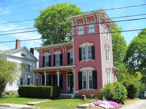 A pink Victorian-style house with black shutters, surrounded by greenery and flowers, under a clear blue sky.