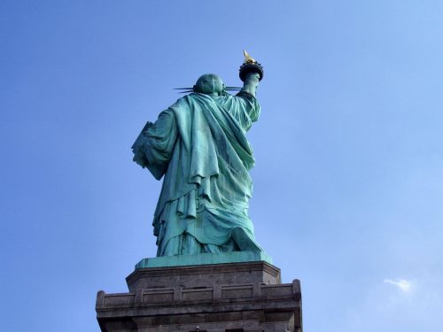 The Statue of Liberty stands tall against a blue sky, holding a torch and facing away from the viewer.