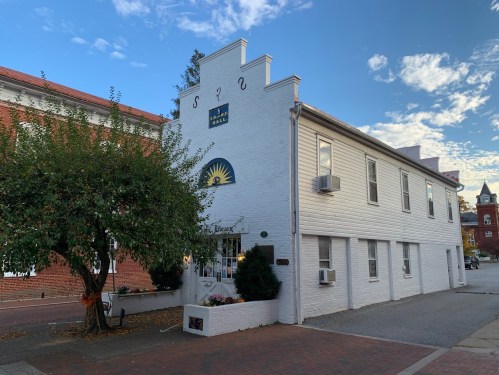 A white building with a sun mural and greenery, located on a brick street with a blue sky in the background.