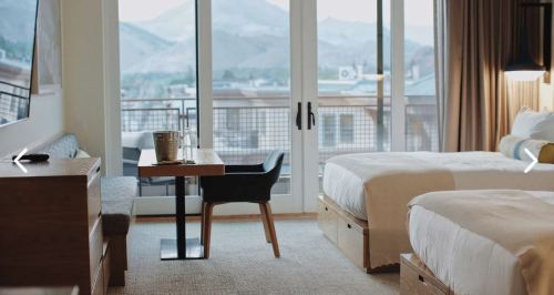 A modern hotel room with two beds, a table, and a balcony view of mountains. Warm lighting and stylish decor.