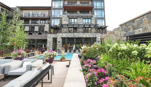 Modern building with stone facade, surrounded by vibrant flowers and greenery, featuring a pool and outdoor seating area.
