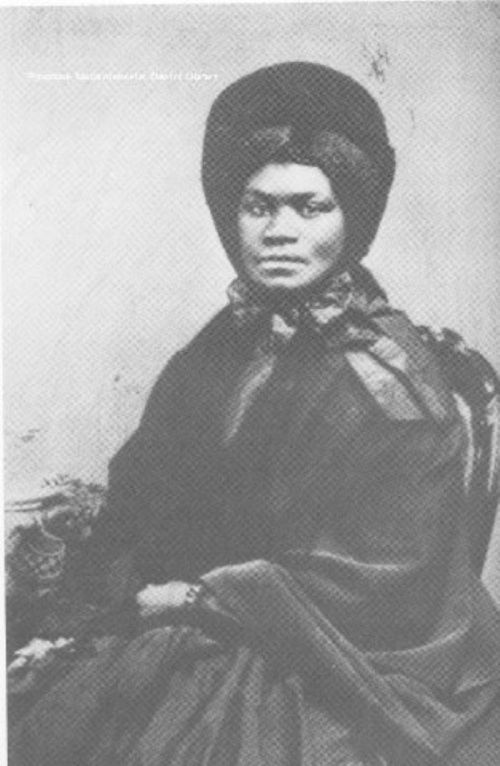 A historical black-and-white portrait of a woman wearing a dark dress and a large hat, seated and looking directly at the camera.
