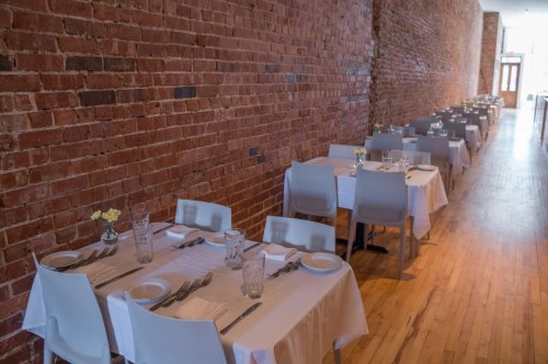 A spacious restaurant with wooden floors, brick walls, and neatly set tables for dining.