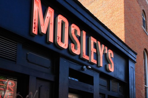 Sign for "Mosley's" in bright neon letters on a dark building exterior.