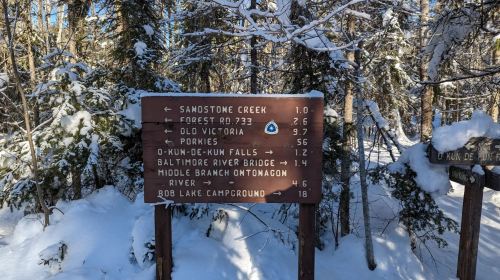 A wooden sign in a snowy forest displaying trail distances to various locations, including Sandstone Creek and Baltimore River Bridge.