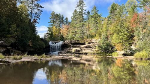 A serene landscape featuring a waterfall surrounded by trees, reflecting in a calm pool of water under a blue sky.