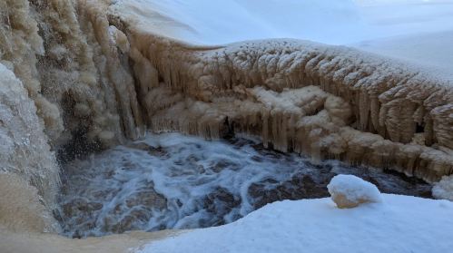 A frozen waterfall with icy formations and flowing water, surrounded by snow-covered terrain.