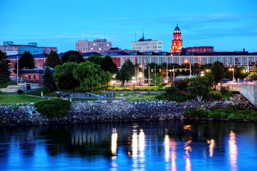 A serene river scene at dusk, reflecting city lights and buildings, with lush greenery along the shore.