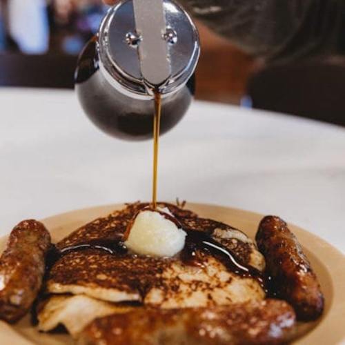 A plate of pancakes topped with butter and syrup, accompanied by two sausage links.