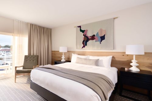 A modern hotel room featuring a bed, decorative wall hanging, and stylish furniture with natural light from a window.