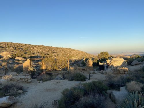 Desert landscape with rocky terrain, sparse vegetation, and remnants of an old structure under a clear blue sky.