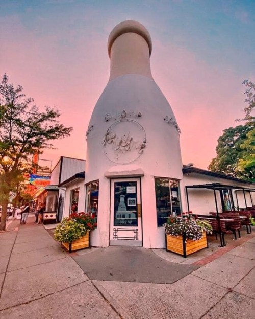 A whimsical building shaped like a milk bottle, surrounded by flowers and trees, under a colorful sunset sky.