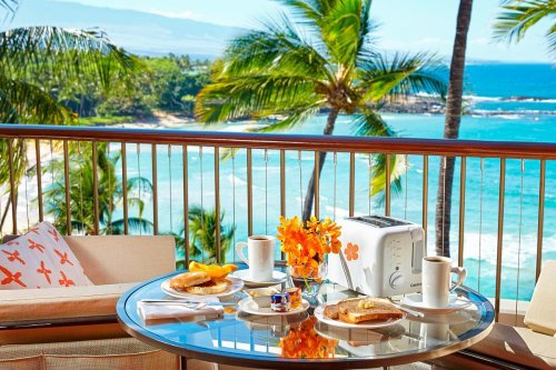 A breakfast table on a balcony overlooking the ocean, featuring toast, coffee, and tropical flowers.