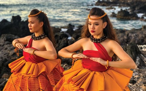 Two women in traditional Hawaiian attire perform a dance by the ocean, surrounded by rocky terrain and water.