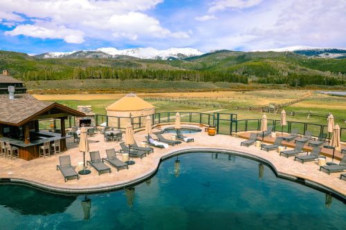 A scenic pool area with lounge chairs, surrounded by mountains and greenery under a partly cloudy sky.