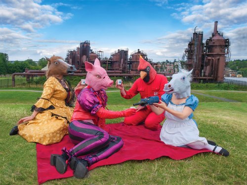 Four people in animal masks (horse, pig, cardinal, unicorn) have a picnic on a red blanket in front of industrial buildings.