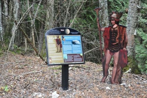 A sign in a forest with information, alongside a life-sized cutout of a historical figure in traditional attire.