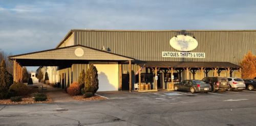 A large metal building with a sign reading "Antiques, Thrift & More," surrounded by trees and parked cars.