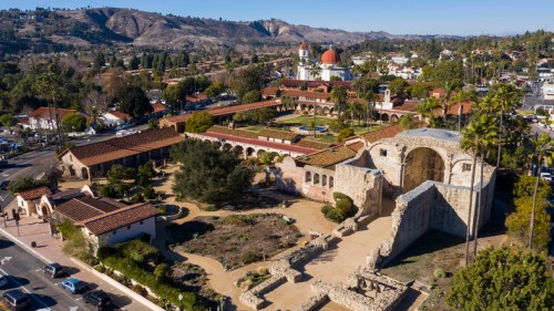 Aerial view of a historic mission complex surrounded by gardens, palm trees, and mountains in the background.