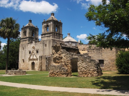 Historic stone ruins of a mission with two bell towers, surrounded by green grass and trees under a blue sky.